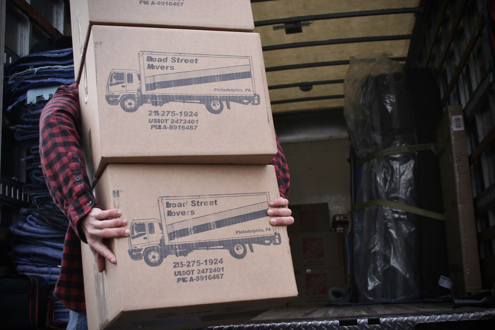 A professional mover carrying three large boxes with the Broad Street Movers logo all at once just before loading them in moving truck